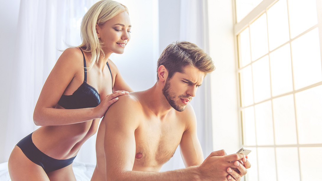 Junges, halbnacktes Paar auf Bett, beide schauen auf ein Handy - Foto: IMAGO / agefotostock