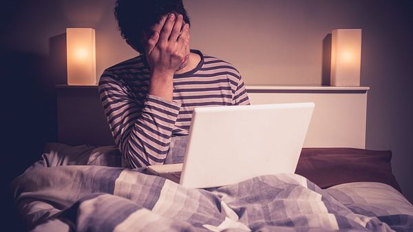 Mann sitzt vor dem Computer im Bett und schlägt die Hand vors Gesicht - Foto: iStock / lolostock