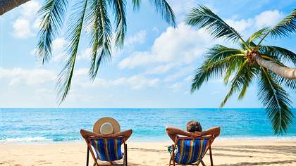 Zwei Urlauber am Strand unter Palmen in Liegestühlen - Foto: IMAGO / Stefan Zeitz
