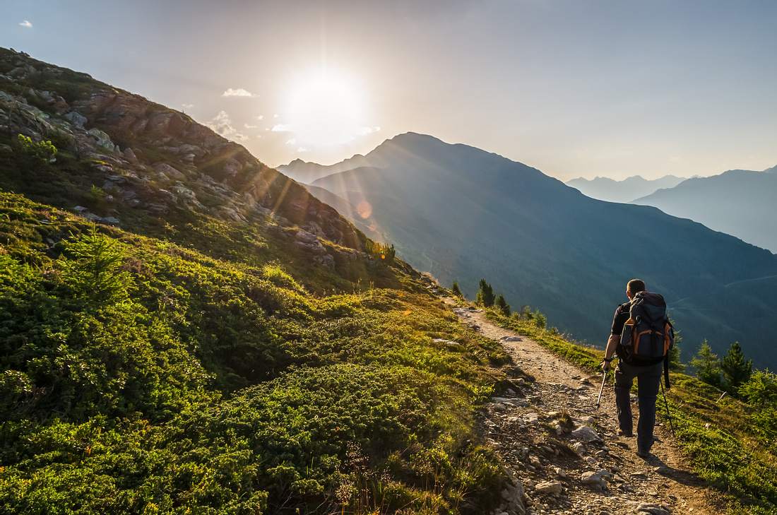 Der richtige Rucksack ist das A&O für einen gelungenen Wandertrip! Ein Mann mit Rucksack wandert bei Sonnenschein durch eine Berglandschaft